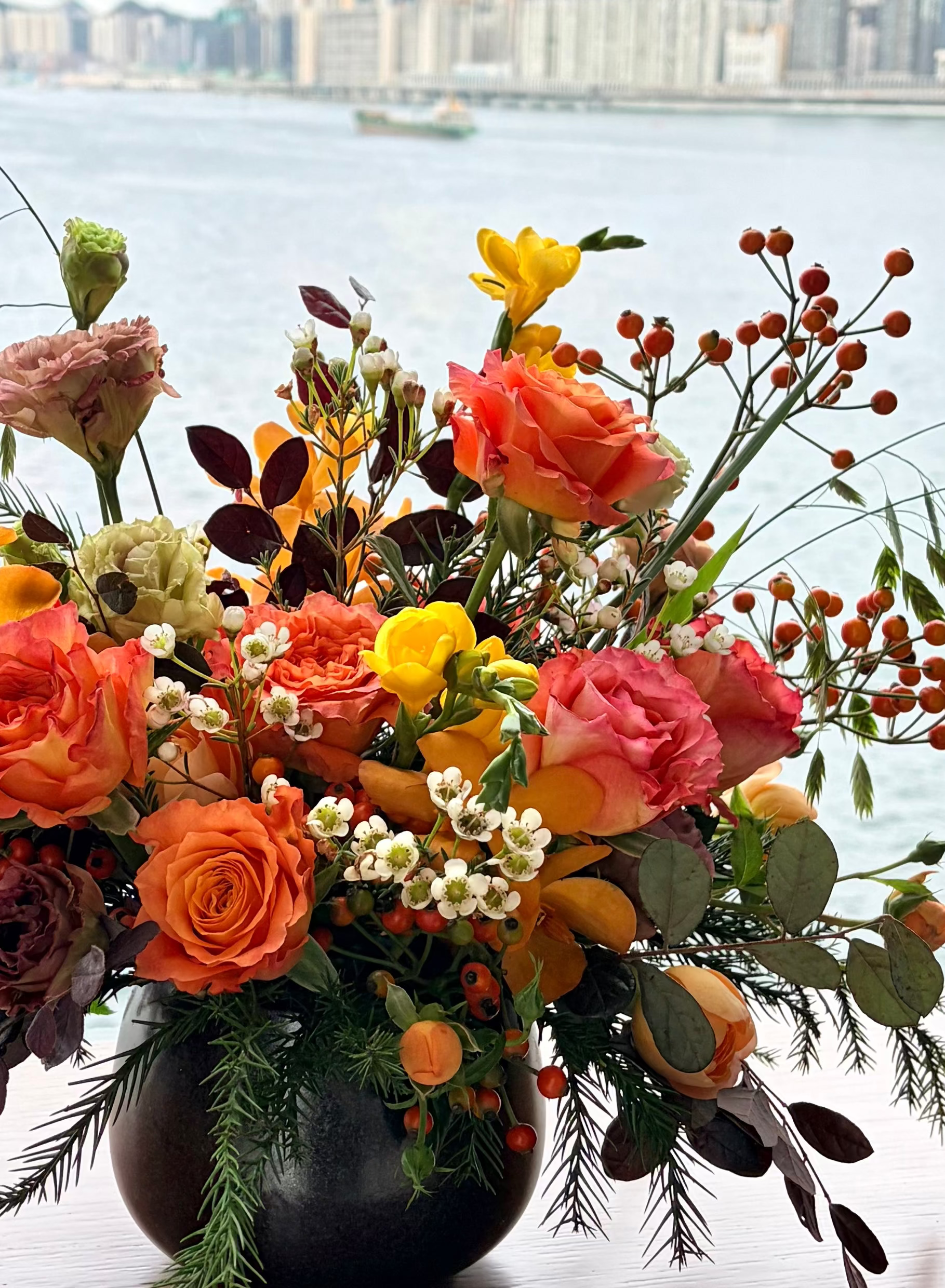 An autumnal floral arrangement in a dark vase, featuring orange and pink roses, yellow flowers, and green foliage, displayed on a wooden ledge with a scenic water and city skyline in the background.